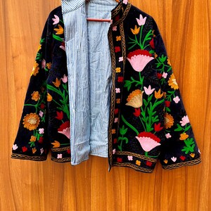 May include: A black velvet jacket with colorful floral embroidery. The jacket has a striped blue and white lining and is hanging on a wooden hanger. The embroidery features pink, orange, and green flowers.