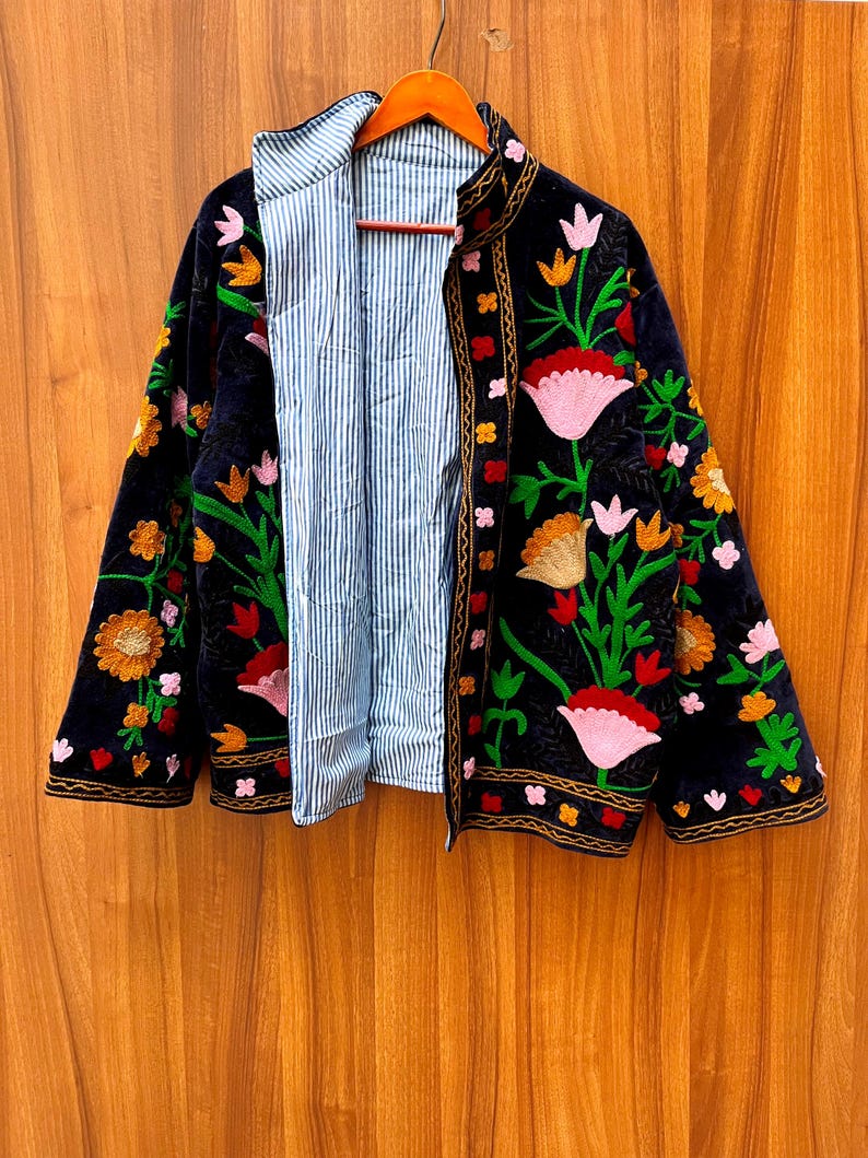 May include: A black velvet jacket with colorful floral embroidery. The jacket has a striped blue and white lining and is hanging on a wooden hanger. The embroidery features pink, orange, and green flowers.