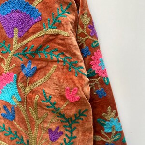May include: Close-up of a rust-colored jacket with intricate floral embroidery. The embroidery features vibrant colors including blue, purple, pink, and green. The jacket's design includes detailed floral patterns and stems.