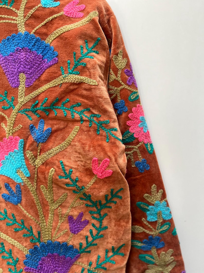 May include: Close-up of a rust-colored jacket with intricate floral embroidery. The embroidery features vibrant colors including blue, purple, pink, and green. The jacket's design includes detailed floral patterns and stems.