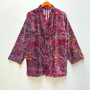 May include: A burgundy paisley patterned jacket with a red and white striped interior. The jacket is made of a soft, textured fabric and has three-quarter length sleeves. The jacket is hanging on a wooden hanger.