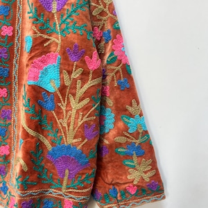 May include: A rust-colored velvet jacket with intricate floral embroidery. The embroidery features vibrant colors including blue, purple, pink, and green. The jacket's design includes detailed floral patterns on the sleeves and front, creating a rich, textured look.