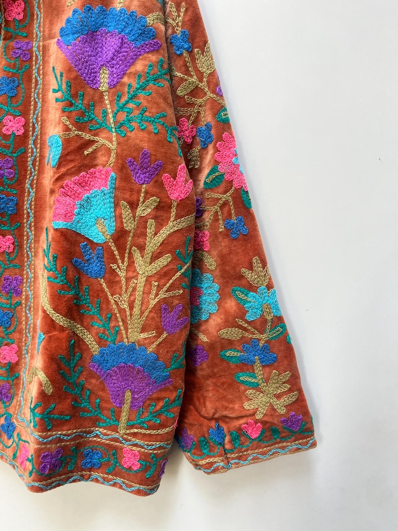 May include: A rust-colored velvet jacket with intricate floral embroidery. The embroidery features vibrant colors including blue, purple, pink, and green. The jacket's design includes detailed floral patterns on the sleeves and front, creating a rich, textured look.