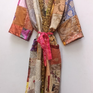 Puede incluir: Una larga bata de retazos con una faja colorida. La bata está hecha de varias telas en tonos marrones, beige, naranja, amarillo, rosa y azul. La faja es de color rosa y naranja brillante.