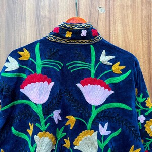 May include: A dark blue velvet jacket with colorful floral embroidery. The jacket features a high collar and is adorned with embroidered flowers in shades of red, yellow, white, and green. The back of the jacket is the focal point, showcasing a large floral design.