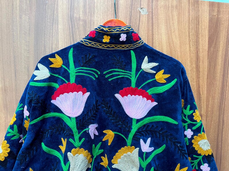 May include: A dark blue velvet jacket with colorful floral embroidery. The jacket features a high collar and is adorned with embroidered flowers in shades of red, yellow, white, and green. The back of the jacket is the focal point, showcasing a large floral design.