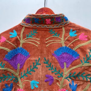 May include: Burnt orange velvet jacket with embroidered floral design. The jacket features a mandarin collar and is adorned with teal, purple, pink, and gold-colored embroidery. The floral pattern is symmetrical and covers the back of the jacket.