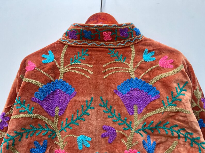 May include: Burnt orange velvet jacket with embroidered floral design. The jacket features a mandarin collar and is adorned with teal, purple, pink, and gold-colored embroidery. The floral pattern is symmetrical and covers the back of the jacket.