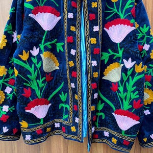 May include: A dark blue embroidered jacket with a floral design. The jacket features red, pink, yellow, and white flowers with green leaves. The edges are trimmed with gold and red embroidery.