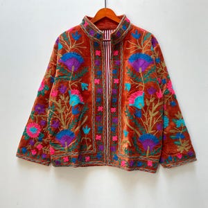 May include: A rust-colored, embroidered jacket with a high collar. The jacket features a floral design in shades of blue, purple, pink, and gold. The jacket is hanging on a wooden hanger.