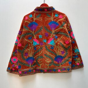 May include: Burnt orange velvet jacket with intricate floral embroidery in shades of purple, blue, pink, and green. The jacket features a mandarin collar and long sleeves, with detailed embroidery along the edges.