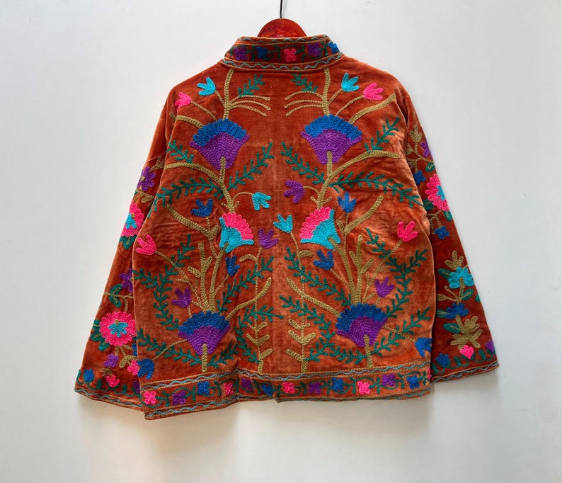 May include: Burnt orange velvet jacket with intricate floral embroidery in shades of purple, blue, pink, and green. The jacket features a mandarin collar and long sleeves, with detailed embroidery along the edges.