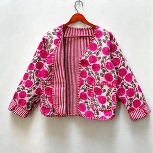 Peut inclure: Une veste matelassée rose et blanche avec un imprimé floral et une doublure rayée. La veste a un col en V et des manches longues.