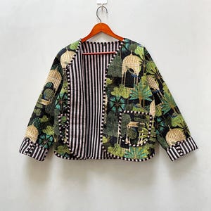 Puede incluir: Una chaqueta acolchada verde y negra con forro a rayas. La chaqueta tiene cuello en V y un bolsillo en el lado izquierdo. La tela presenta un estampado floral con pájaros.