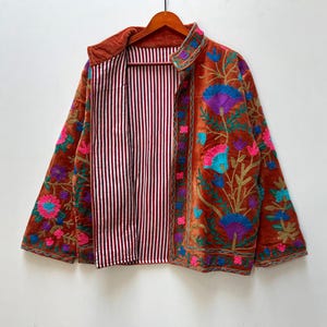 May include: A rust-colored, open-front jacket with intricate floral embroidery in shades of blue, purple, pink, and green. The jacket's interior features a striped pattern in burgundy and white. The jacket is hanging on a wooden hanger.