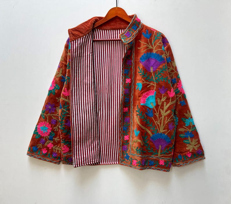 May include: A rust-colored, open-front jacket with intricate floral embroidery in shades of blue, purple, pink, and green. The jacket's interior features a striped pattern in burgundy and white. The jacket is hanging on a wooden hanger.