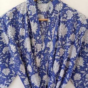 Op de afbeelding: Een blauwe en witte katoenen badjas met een bloemenprint en witte bies. De badjas heeft een strikceintuur en lange mouwen.