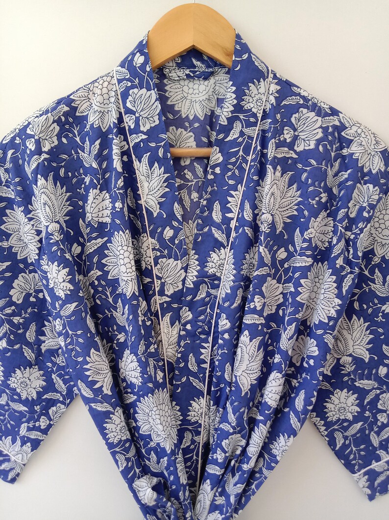 Op de afbeelding: Een blauwe en witte katoenen badjas met een bloemenprint en witte bies. De badjas heeft een strikceintuur en lange mouwen.