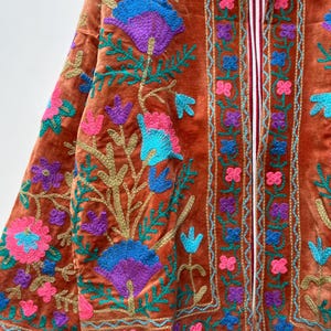 May include: Rust-colored velvet jacket with intricate floral embroidery in shades of blue, purple, and pink. The jacket features a striped lining and gold-colored thread detailing, creating a rich, textured look.