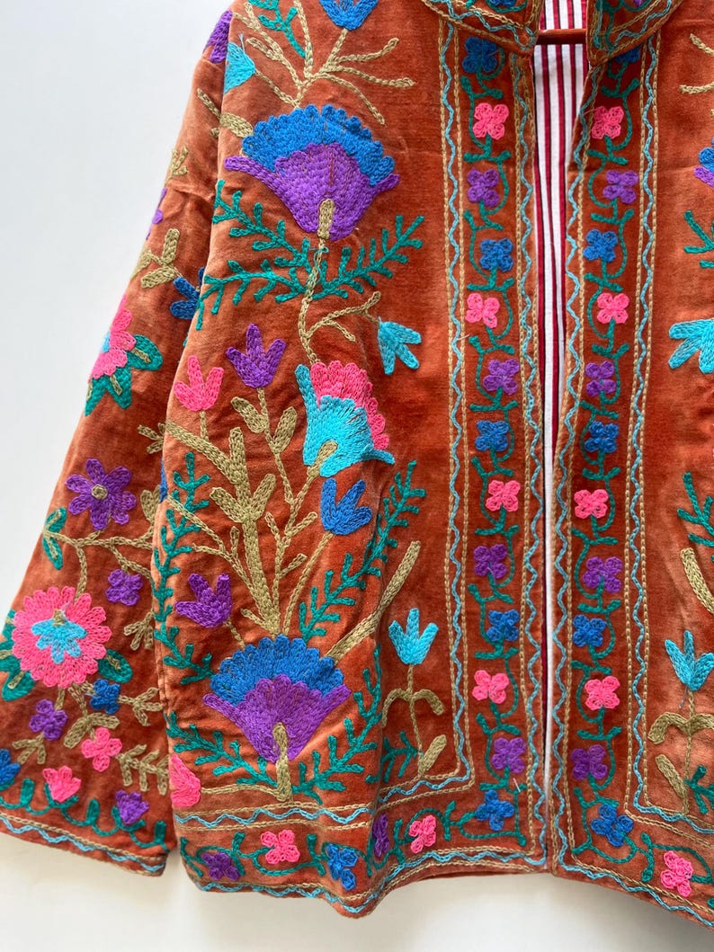 May include: Rust-colored velvet jacket with intricate floral embroidery in shades of blue, purple, and pink. The jacket features a striped lining and gold-colored thread detailing, creating a rich, textured look.