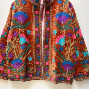 May include: A rust-colored velvet jacket with intricate floral embroidery in shades of blue, purple, pink, and gold. The jacket features a striped lining and a collarless design, showcasing a bohemian aesthetic.