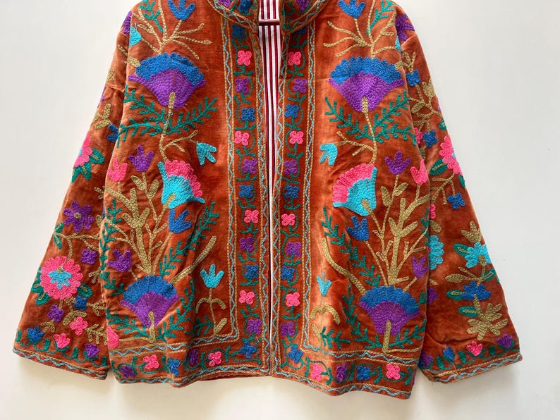 May include: A rust-colored velvet jacket with intricate floral embroidery in shades of blue, purple, pink, and gold. The jacket features a striped lining and a collarless design, showcasing a bohemian aesthetic.