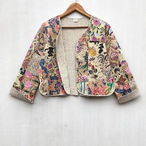 May include: A quilted jacket with a floral pattern in shades of pink, blue, and yellow. The jacket has a striped lining and is hanging on a wooden hanger. The jacket's sleeves are three-quarter length.