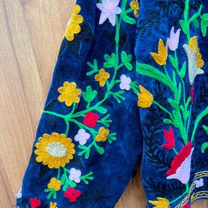 May include: Close-up of a navy blue embroidered jacket sleeve. The jacket features colorful floral embroidery in yellow, pink, red, and green. The edges are trimmed with gold thread, creating a detailed and ornate design. The jacket is made of a soft, velvet-like material.