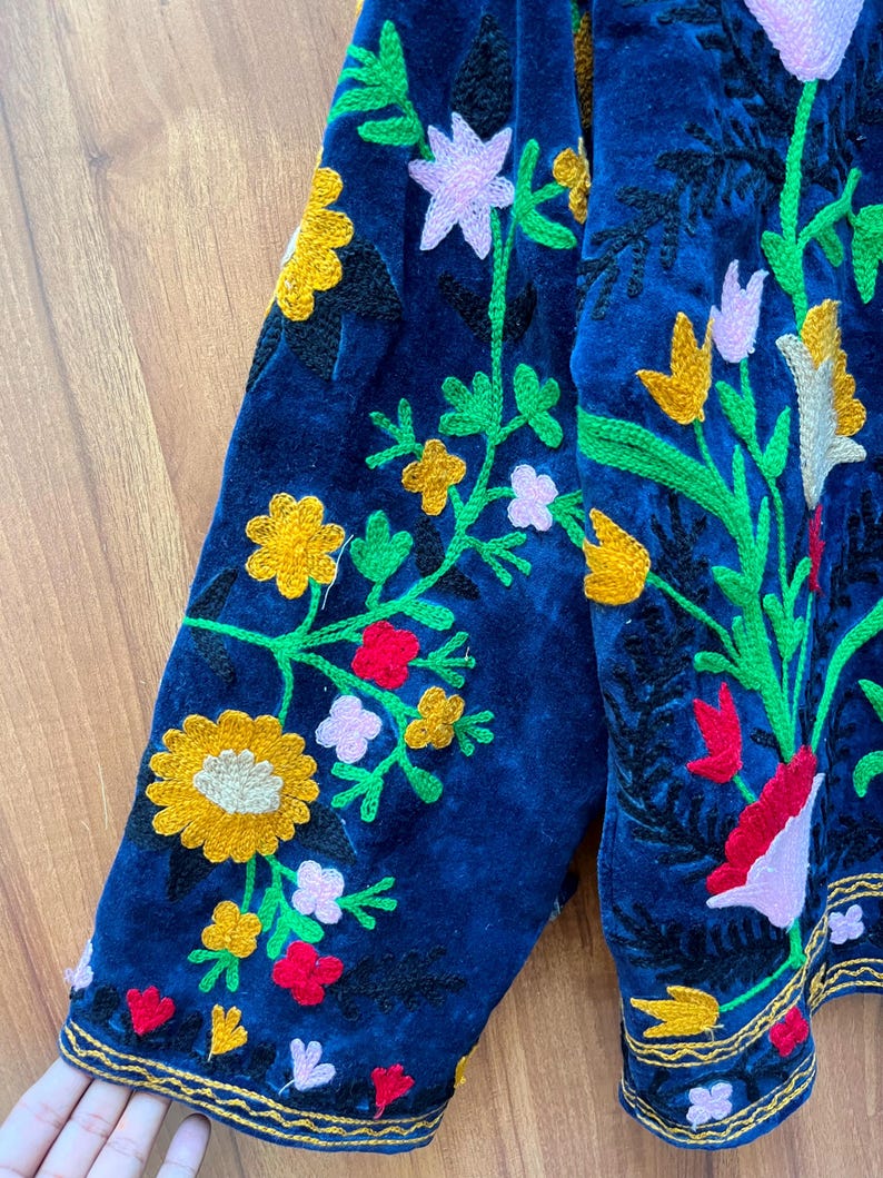 May include: Close-up of a navy blue embroidered jacket sleeve. The jacket features colorful floral embroidery in yellow, pink, red, and green. The edges are trimmed with gold thread, creating a detailed and ornate design. The jacket is made of a soft, velvet-like material.
