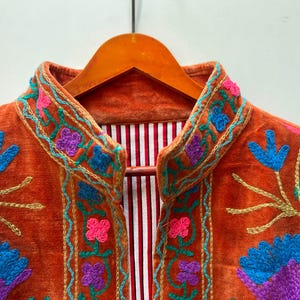 May include: Close-up of an orange velvet jacket with colorful floral embroidery. The jacket features blue, purple, pink, and gold thread detailing. The interior lining has a red and white striped pattern.
