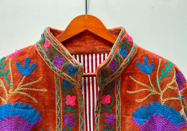May include: Close-up of an orange velvet jacket with colorful floral embroidery. The jacket features blue, purple, pink, and gold thread detailing. The interior lining has a red and white striped pattern.