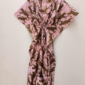 May include: A pink and brown patterned kaftan dress hanging on a wooden hanger. The dress features a repeating pattern of brown cheetahs on a pink background with small white dots. The kaftan has a V-neck and a tie at the waist.