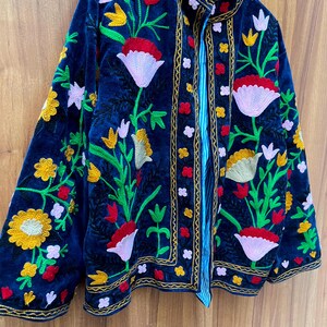 May include: A navy blue velvet jacket with colorful floral embroidery. The jacket features a high collar and is adorned with red, yellow, and white flowers, along with green leaves. The edges are trimmed with gold stitching, and the lining is striped.
