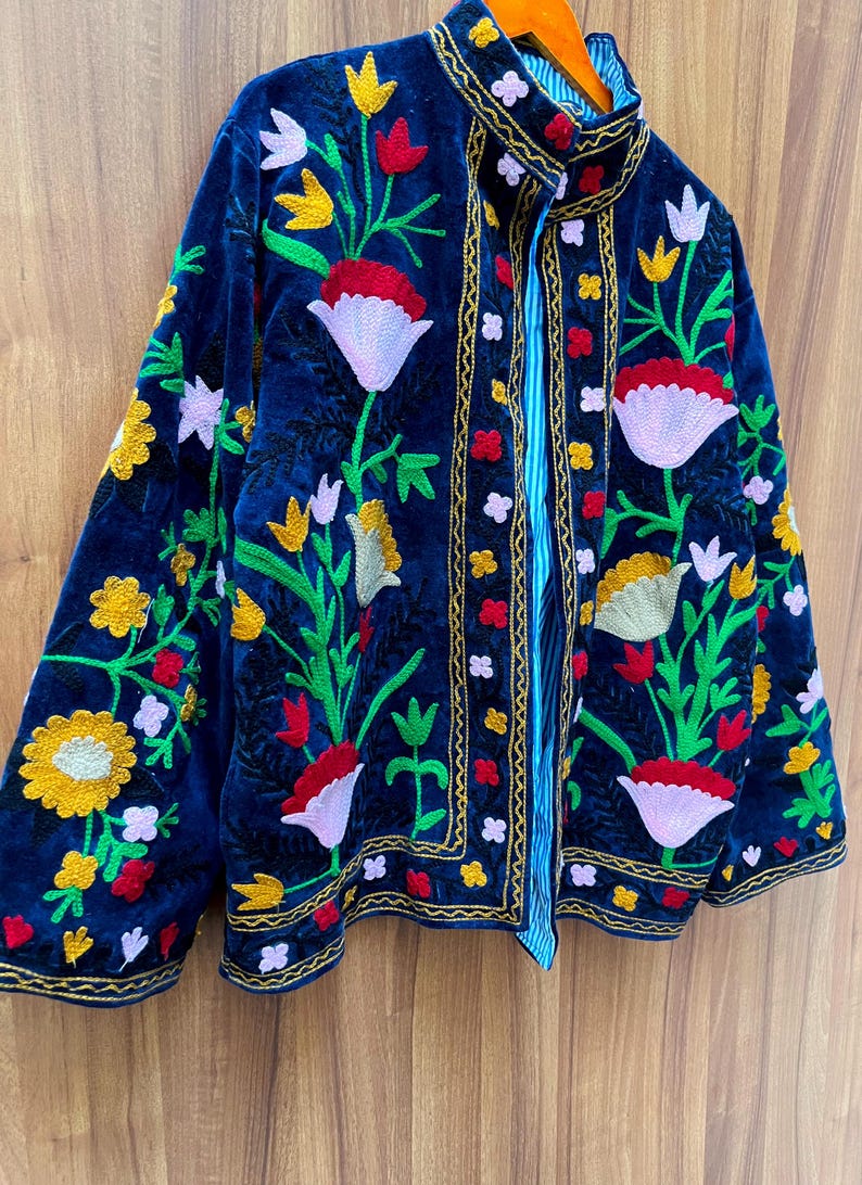 May include: A navy blue velvet jacket with colorful floral embroidery. The jacket features a high collar and is adorned with red, yellow, and white flowers, along with green leaves. The edges are trimmed with gold stitching, and the lining is striped.