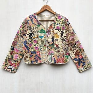 Könnte beinhalten: Beige gesteppte Jacke mit V-Ausschnitt und langen Ärmeln. Die Jacke hat ein Patchwork-Design mit Blumen- und Pflanzenmustern in Rosa-, Blau- und Grüntönen. Die Jacke hängt an einem Holzbügel.