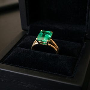 Peut inclure: Une bague en or avec une grosse pierre verte taillée en émeraude, sertie dans un anneau en or. La bague est présentée dans un écrin noir doublé de velours avec un couvercle à charnière. La boîte est en bois foncé.