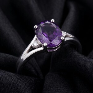 May include: A silver ring featuring a large, oval-cut purple amethyst gemstone. Two smaller, round, clear gemstones flank the main stone. The ring is set against a dark background, highlighting the ring's design.