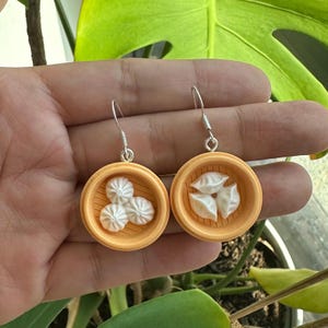 Op de afbeelding: Een paar zilveren oorhangers met een wit keramisch knoedelontwerp. De knoedels bevinden zich in een klein, rond, oranje mandje.
