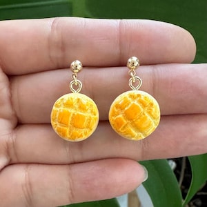 May include: A pair of gold earrings with a yellow and white bread-shaped charm. The charms have a textured surface that resembles a pastry.