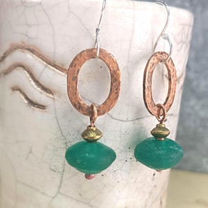 Könnte beinhalten: Ein Paar handgefertigte Ohrringe mit gehämmerten ovalen Kupferreifen, silbernen Ohrhaken und tropfenförmigen grünen Perlen. Die Ohrringe haben kleine goldfarbene Akzente und eine rosa Perle am unteren Ende der grünen Perle.