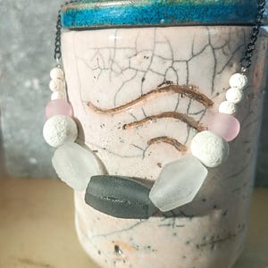 Puede incluir: Un collar artesanal con una cadena negra y una variedad de cuentas. El collar incluye cuentas blancas, rosas y grises, así como cuentas transparentes de forma geométrica. El collar se muestra sobre un fondo rosa y azul.