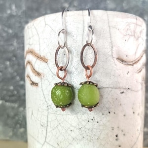 Puede incluir: Un par de pendientes hechos a mano con un diseño único. Los pendientes presentan ganchos para las orejas de color plateado, eslabones ovalados de color cobre y pequeñas esferas de cuentas verdes. Las cuentas están acentuadas con tapas de color cobre.