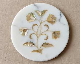 May include: Round white marble coaster with an inlaid floral design. The design features a central stem with leaves and flowers in shades of gold and mother-of-pearl. The coaster is approximately 4 inches in diameter.