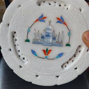 May include: White marble coaster with an inlaid Taj Mahal design. The coaster features a detailed depiction of the iconic monument in blue and white, surrounded by floral accents in red, blue, and green. The outer edge has a carved leaf pattern.