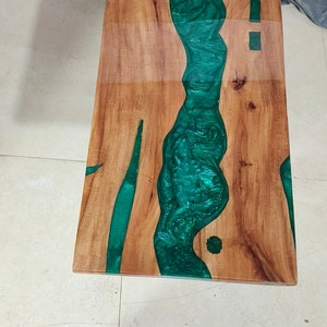 May include: A rectangular wooden table with a river of green epoxy resin running through the center. The wood has a natural grain, and the resin has a glossy finish. The table is a unique piece of furniture.