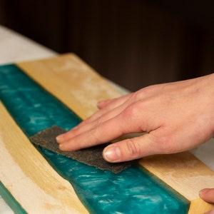 May include: A close-up of a wooden board with a teal epoxy resin river. A hand is sanding the surface with a piece of sandpaper. The wood is light-colored, and the resin has a swirling, ocean-like appearance.