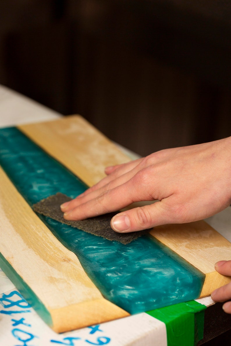 May include: A close-up of a wooden board with a teal epoxy resin river. A hand is sanding the surface with a piece of sandpaper. The wood is light-colored, and the resin has a swirling, ocean-like appearance.