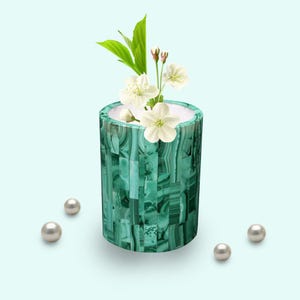 May include: A cylindrical, green malachite vase with a white interior, holding white flowers and green leaves. Four pearls are scattered around the vase on a light blue background. The vase has a mosaic-like pattern.