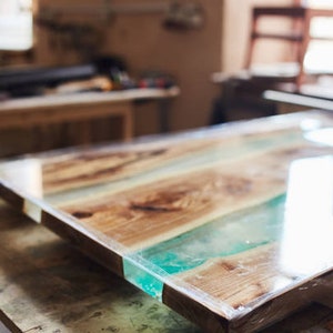 May include: A rectangular wooden table with a clear epoxy resin finish. The table features alternating wooden planks and sections of turquoise resin. The table is in a workshop setting, with other wooden furniture visible in the background.