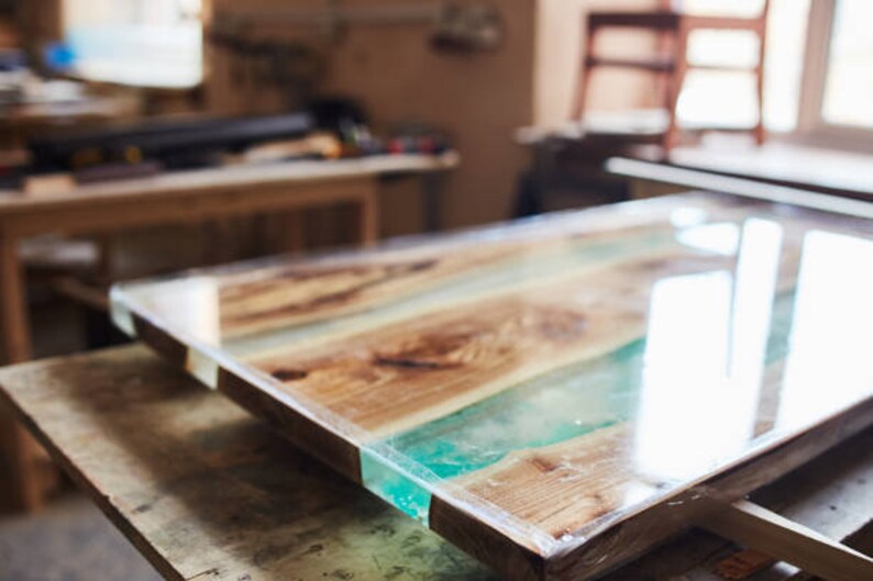 May include: A rectangular wooden table with a clear epoxy resin finish. The table features alternating wooden planks and sections of turquoise resin. The table is in a workshop setting, with other wooden furniture visible in the background.